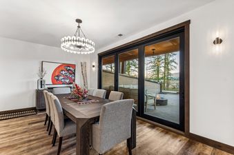 A modern dining room with a long wooden table and upholstered chairs under a circular chandelier, with sliding glass doors opening to an outdoor patio with a fire pit.
