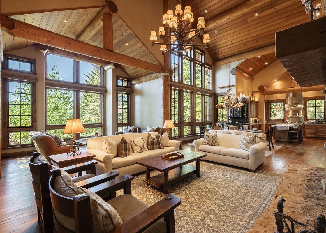 The Deer Valley Oak Lodge features a vast living area with high ceilings, exposed wooden beams, and large windows that offer views of lush greenery.