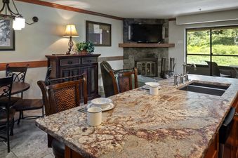 An inviting open-concept living and dining area at Building 4 One Bedroom Beaver Run Resort, featuring a granite kitchen island, comfortable seating, a stone fireplace, and a large window overlooking a lush outdoor landscape.