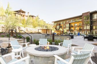 An inviting outdoor fire pit area at Lift 406 with a lit fire pit surrounded by Adirondack chairs, set against a modern building and green hillside.