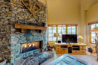 A cozy living area features a stone fireplace with a roaring fire, a flat-screen TV on a wooden entertainment center, and rustic decor, including snowshoes and pinecones.