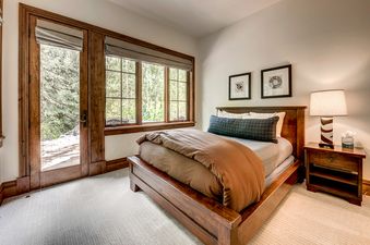 A cozy bedroom with a wooden bed frame, brown bedding, and a view of the forest outside the window.