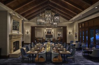A spacious, warmly lit dining room with several long tables set for dinner, featuring a large stone fireplace and large chandeliers.