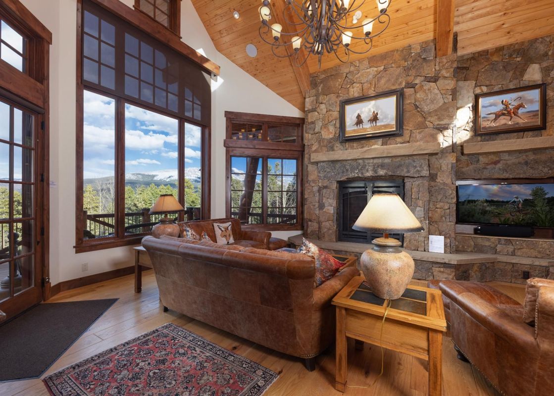 The living room at Fox Paw Lodge features a large fireplace made of stacked stone, comfortable leather sofas, and expansive windows that offer stunning views of the snow-capped mountains and lush forest.