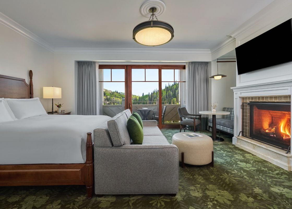 The Montage Deer Valley King Room Vista View features a king-sized bed with white linens, a comfortable seating area, a fireplace, and a private balcony overlooking a lush, forested mountain landscape.