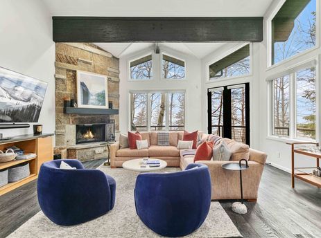 A modern, bright living room in Twin Pines 6 featuring a large stone fireplace, cozy sectional, and two blue velvet armchairs with expansive windows looking out at the trees.