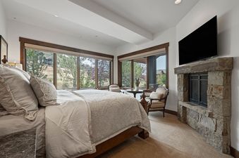 A luxurious master bedroom at Juniper Landing 108 features a large bed, a stone fireplace with a TV, and panoramic windows with outdoor views.