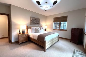 A cozy bedroom with a king-sized bed, two nightstands with lamps, a window with blinds, and a brown dresser in the corner.