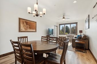 The open-concept dining and living area of Columbia Crest 301 features a round wooden dining table with six chairs, a comfortable sofa, armchairs, and a TV, with large windows overlooking trees.