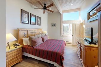 Bedroom in Manor Vail 364, showcasing a plush bed with red and white bedding, a side table with a lamp and a phone, a window with an exterior view, and a dresser with a flat screen television on top.
