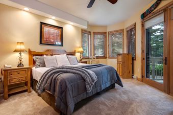 A cozy king bedroom at Chateau Chamonix CX315 features a wooden bed, gray bedding, warm lighting, and windows with wooden blinds looking out onto greenery.