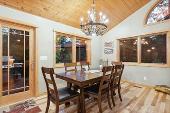 A rustic dining area with a wooden table, chairs, an antler chandelier, and large windows looking out to nature.