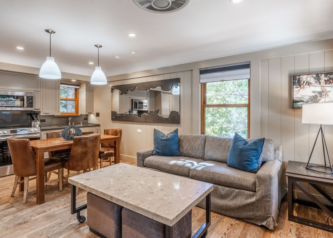 An open concept living area at Cedar Crest Sparrow Cottage 1 featuring a modern kitchen, a dining table with leather chairs, and a comfortable living room with a gray sofa and blue pillows.