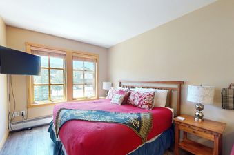 A bedroom with a red bedspread, two windows, and a wooden nightstand.