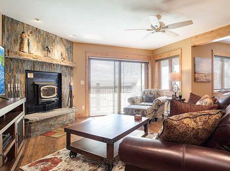 A cozy, rustic living room featuring a stone fireplace, a large flat-screen TV displaying a river scene, and comfortable leather and patterned seating.