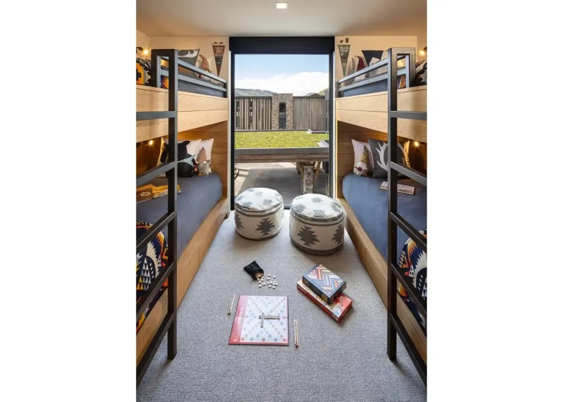 A modern bunk room featuring two sets of wooden and black metal bunk beds with navy bedding and patterned pillows, two round patterned poufs, and a board game on a grey carpet, with a large window offering an outdoor view.