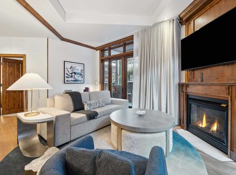The living room of The Aspen 03B features a fireplace with a large TV above it and a large window looking out onto a balcony.