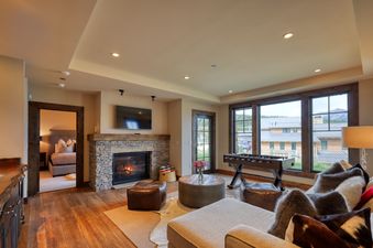 A rustic living room with a stone fireplace, leather ottomans, a foosball table, and large windows revealing a mountain view at Tanager Cabin 43.