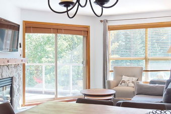 A cozy living room at Aspens on Blackcomb Unit 332 features a stone fireplace, mounted TV, and large windows leading to an exterior view.