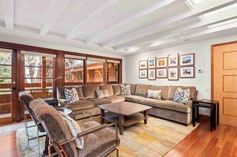 A cozy living room featuring a large, L-shaped sectional sofa with throw pillows, two armchairs with sheepskin throws, a coffee table, and framed art on the wall, with windows revealing a snowy outdoor view.