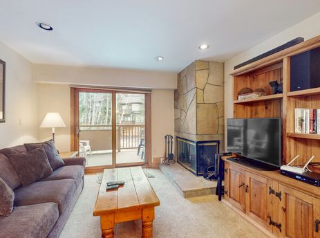 A cozy living room at Fallridge 402 featuring a comfortable sofa, a large stone fireplace, a wooden entertainment center with a TV and books, and a sliding glass door leading to a balcony.
