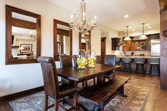A dining area with a rustic wooden table, leather chairs, and a matching bench, featuring decorative vases with fruit, leading into a modern kitchen with bar seating and stainless steel appliances.