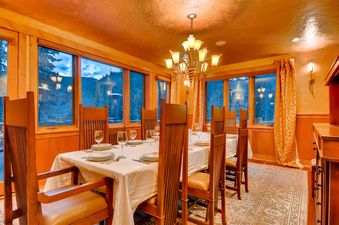 The dining room at Alders Chalet features a large wooden table set for a meal, surrounded by matching wooden chairs, with expansive windows offering a view of the snow-covered trees and mountains.