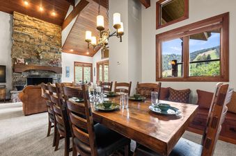 A spacious dining room with a large wooden table set for a meal, surrounded by wooden chairs, leading into a living area with a stone fireplace, all under a vaulted wooden ceiling with views of green mountains through large windows.