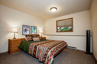 A cozy bedroom in The Cedar Nest features a bed with a patterned comforter, wooden nightstands, lamps, framed artwork, and a window.