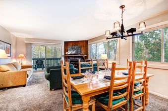 The dining and living area of Snow Flower 150 Park City, featuring a rustic wooden dining table, a cozy living room with a fireplace, and large windows overlooking autumn trees.