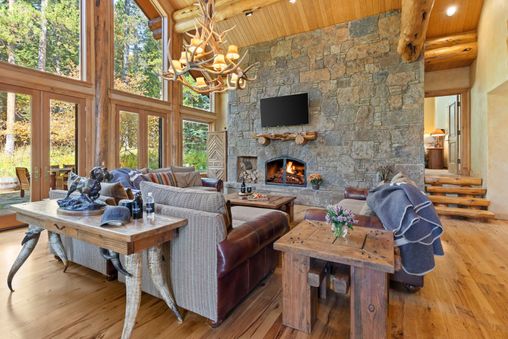 A cozy living room with a stone fireplace, large windows overlooking the forest, and rustic wooden furniture.
