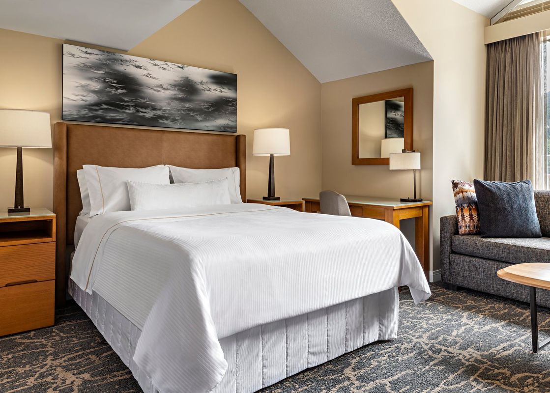 A spacious bedroom in The Westin Resort Deluxe Studio Suite Mountain View features a comfortable bed with white linens, a nightstand with a lamp, a desk with a chair, and a sofa.