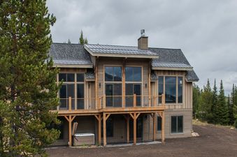 An impressive three-story vacation home, Hunts View, features extensive wooden paneling, a gray shingled roof, a spacious balcony, and a patio, all surrounded by lush pine trees.