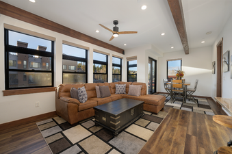 The spacious living area within Arrow Townhome 202 features a large brown sectional sofa, a dark wood coffee table, and a patterned area rug, with a dining area visible in the background.