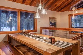 A wooden dining table with a black and white checkered table runner sits in a spacious living room, with a view of a snowy forest outside the window.