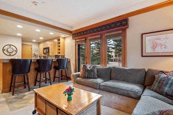 A spacious and inviting open-concept living area with a comfortable L-shaped sofa, a wooden coffee table, and a well-appointed kitchen with a breakfast bar and bar stools.