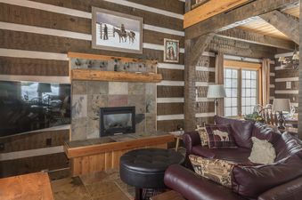 A rustic cabin living room with a fireplace, a large leather sofa with decorative pillows, and wood-paneled walls.