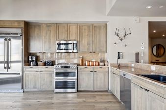 A modern kitchen with light wooden cabinetry, granite countertops, and stainless steel appliances including a refrigerator, microwave, and double oven.
