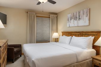 A serene bedroom in Montaneros 312 features a large bed with white linens, a wooden headboard, a bedside lamp, an armchair, and a window with drapes.