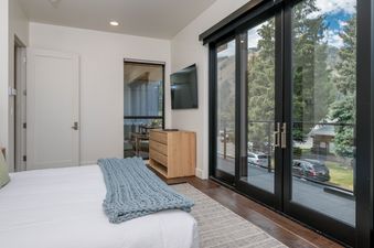 A bedroom with a large bed, a dresser, a television, and a door leading to a balcony overlooking trees.