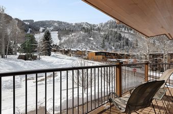 A private balcony with two chairs overlooks a snow-covered mountain landscape with ski slopes and modern buildings.