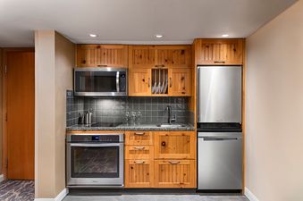 A fully equipped compact kitchen featuring wooden cabinetry, stainless steel appliances, and a grey tiled backsplash.
