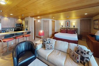 A living room with a white leather couch and a black leather armchair, a bedroom with a white queen bed, and a kitchen with bar stools.