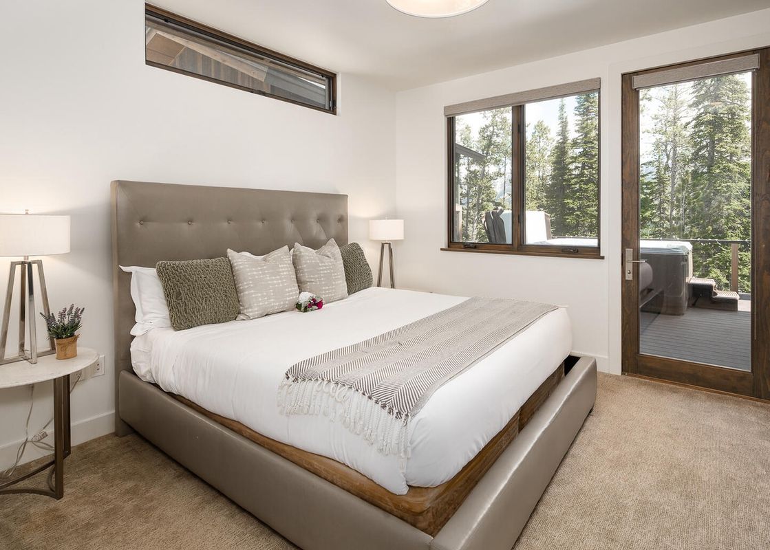 The bedroom at Moonlight Basin 8 Alpine Bend features a king-size bed with white linens and a grey patterned throw, a large window overlooking the trees, and a door leading to a patio with a hot tub.