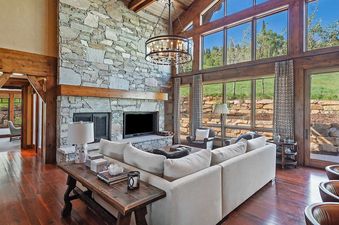 The living room at Solstice at Empire Pass features a large stone fireplace, comfortable white sofas, and floor-to-ceiling windows with expansive views of the outdoors.