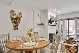 A dining table set for four with a rustic wood table, white plates, and a floral centerpiece in front of a fireplace.