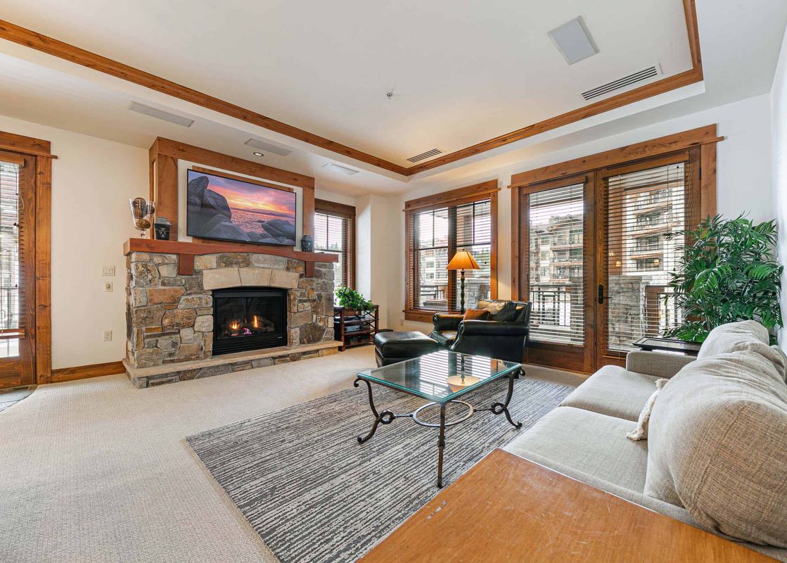 The living room of Big Horn Lodge 202 features a stone fireplace, a flatscreen TV, a large couch, a glass coffee table, a comfy chair, and large windows that offer views of the surrounding area.