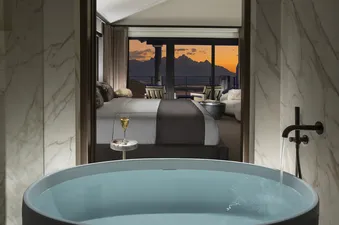 A luxurious bathroom with a freestanding tub filling with water, looking into a bedroom with a king-sized bed and a balcony with mountain views at sunset.