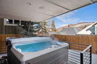 An outdoor hot tub with steaming water is featured on a balcony, offering a stunning view of snow-covered mountains under a clear blue sky.