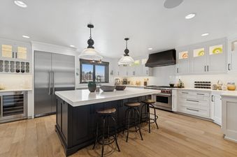 A spacious, modern kitchen with white cabinetry, stainless steel appliances, a large island with three bar stools, and light wood flooring.